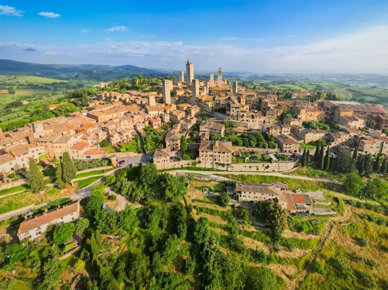 Aerial view of medieval towns in Italy, with ancient towers and terracotta roofs nestled in rolling green hills under a blue sky.