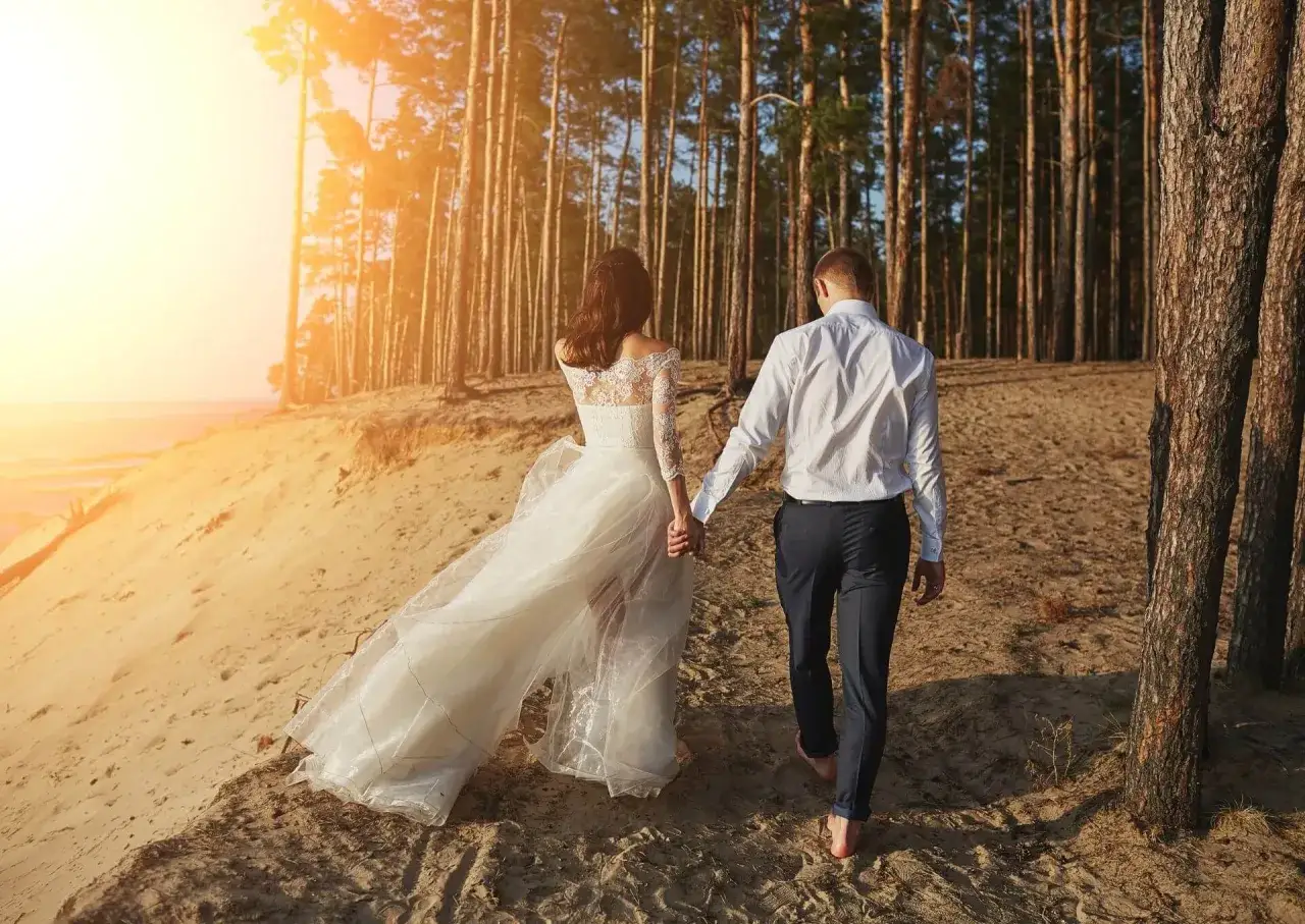 Para młoda idzie boso przez piaszczystą plażę w lesie sosnowym, celebrując swój ślub humanistyczny. Słońce zachodzi, rzucając ciepłe światło.