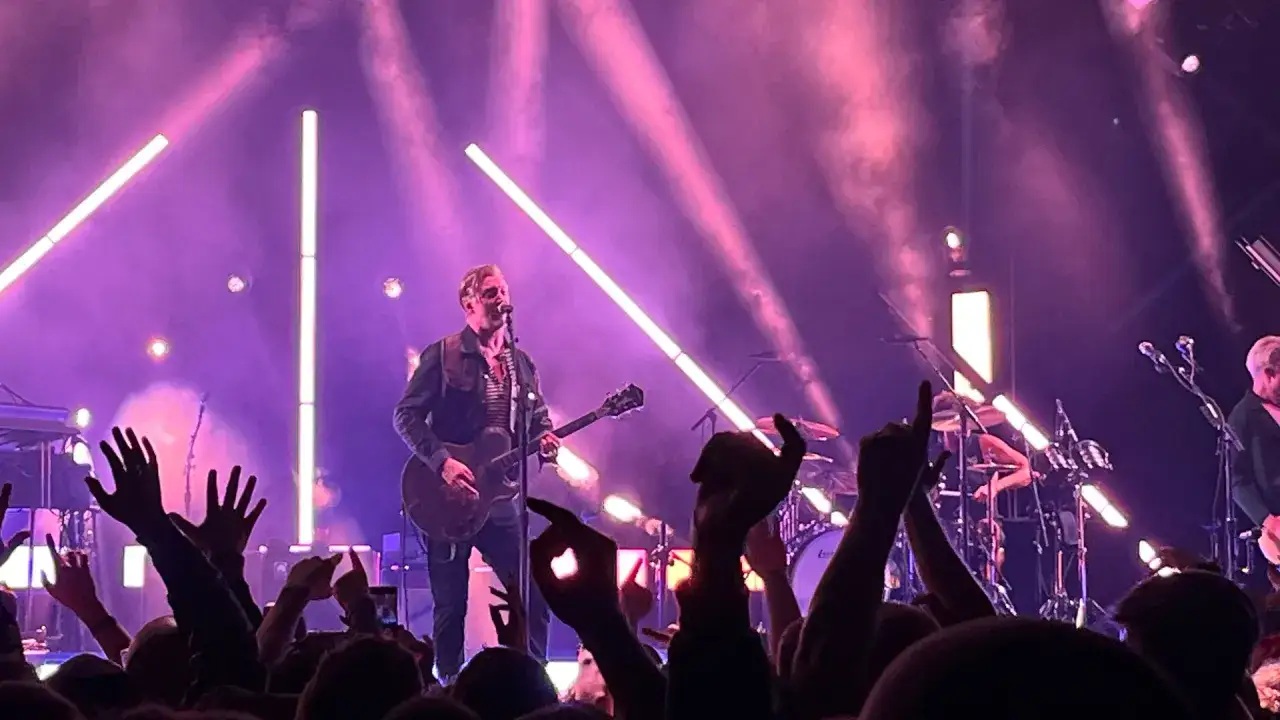 Concert des membres de Queens of the Stone Age sous des &eacute;clairages roses. La foule l&egrave;ve les mains en signe d'enthousiasme.