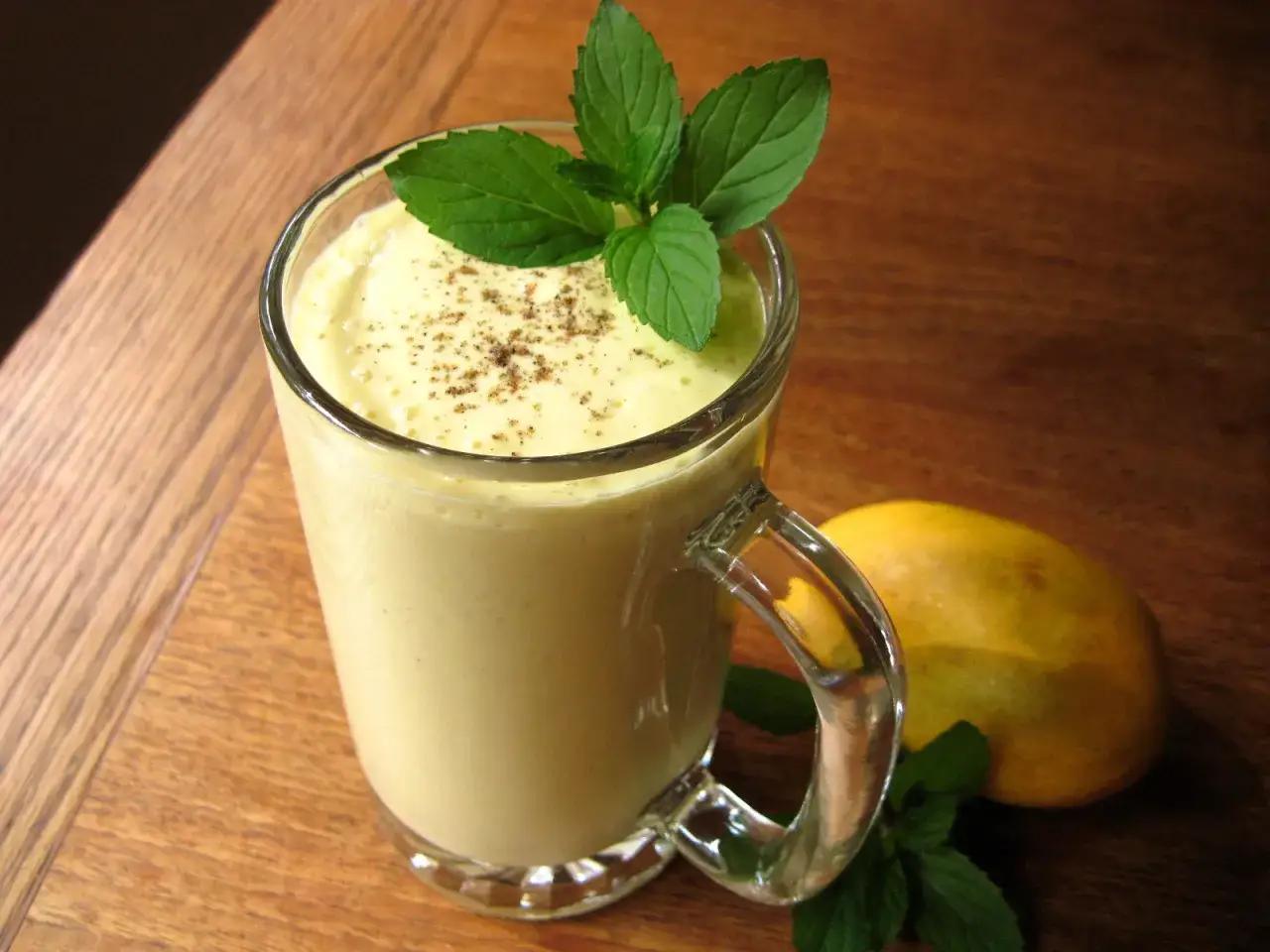 En krämig mango lassi i ett glas, garnerad med mynta och en nypa kryddor. En hel mango ligger bredvid.