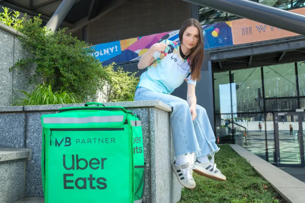 Młoda kobieta w koszulce z logo Uber Eats i jeansach siedzi obok zielonej torby dostawcy.