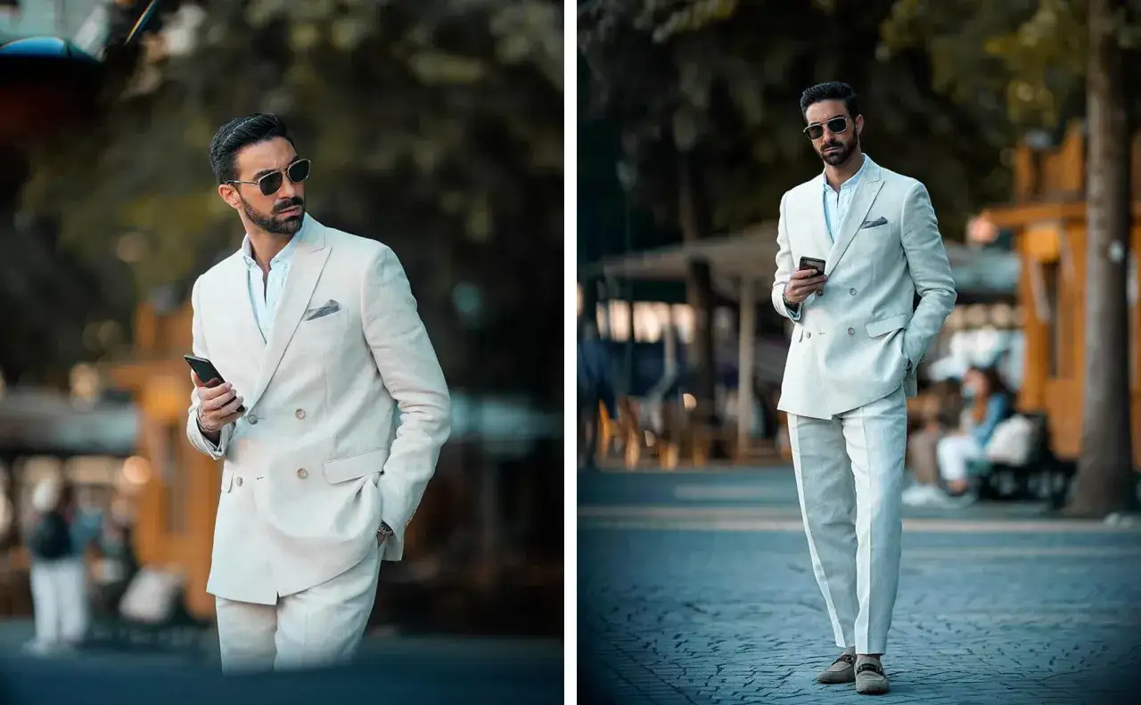 Uomo con abito **semi formal** color crema, occhiali da sole e smartphone in mano, passeggia in una strada acciottolata.