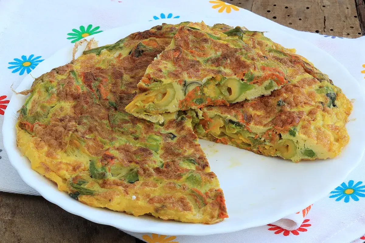 Frittata dorata con fiori di zucca, carote e olive, tagliata a spicchi su un piatto bianco.