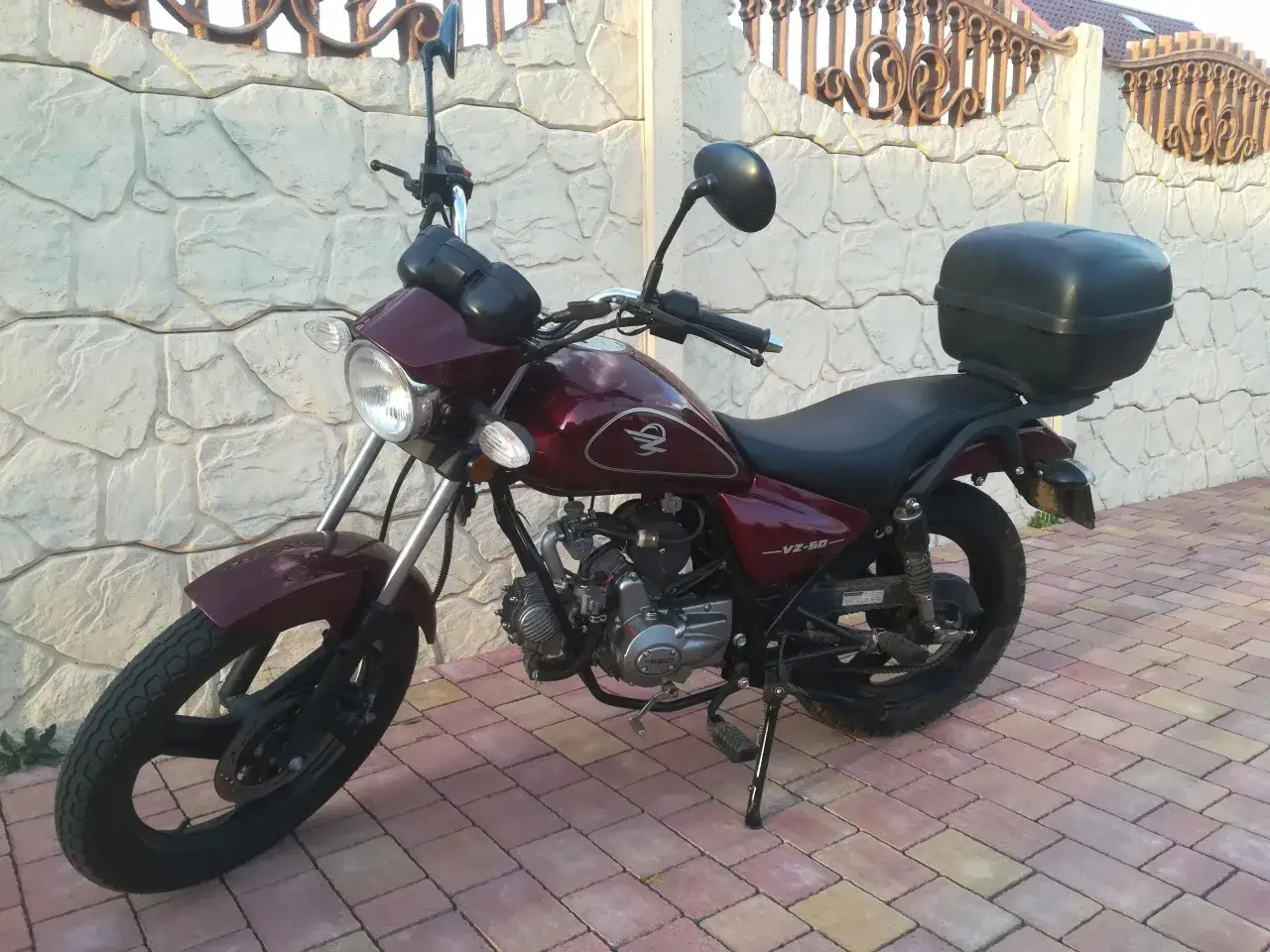 Ciemnoczerwony motorower na dowód osobisty, z czarnym kufrem z tyłu, stoi na wybrukowanym podjeździe przy kamiennym murze.