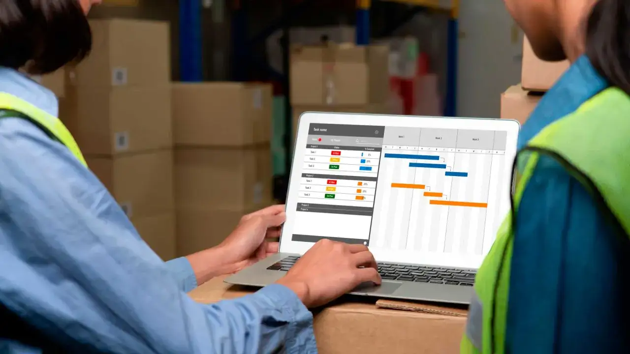 Due operai in un magazzino analizzano un diagramma di Gantt su un laptop, pianificando la richiesta turno fisso.