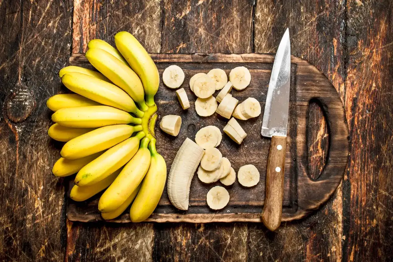 Pęczek banan&oacute;w, pokrojone plasterki i n&oacute;ż na desce. Czy banany można trzymać w lod&oacute;wce? Tutaj dowiesz się, jak najlepiej je przechowywać.