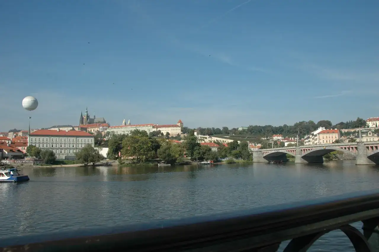 Widok na rzekę w Pradze z balonem unoszącym się nad miastem i mostem w oddali.