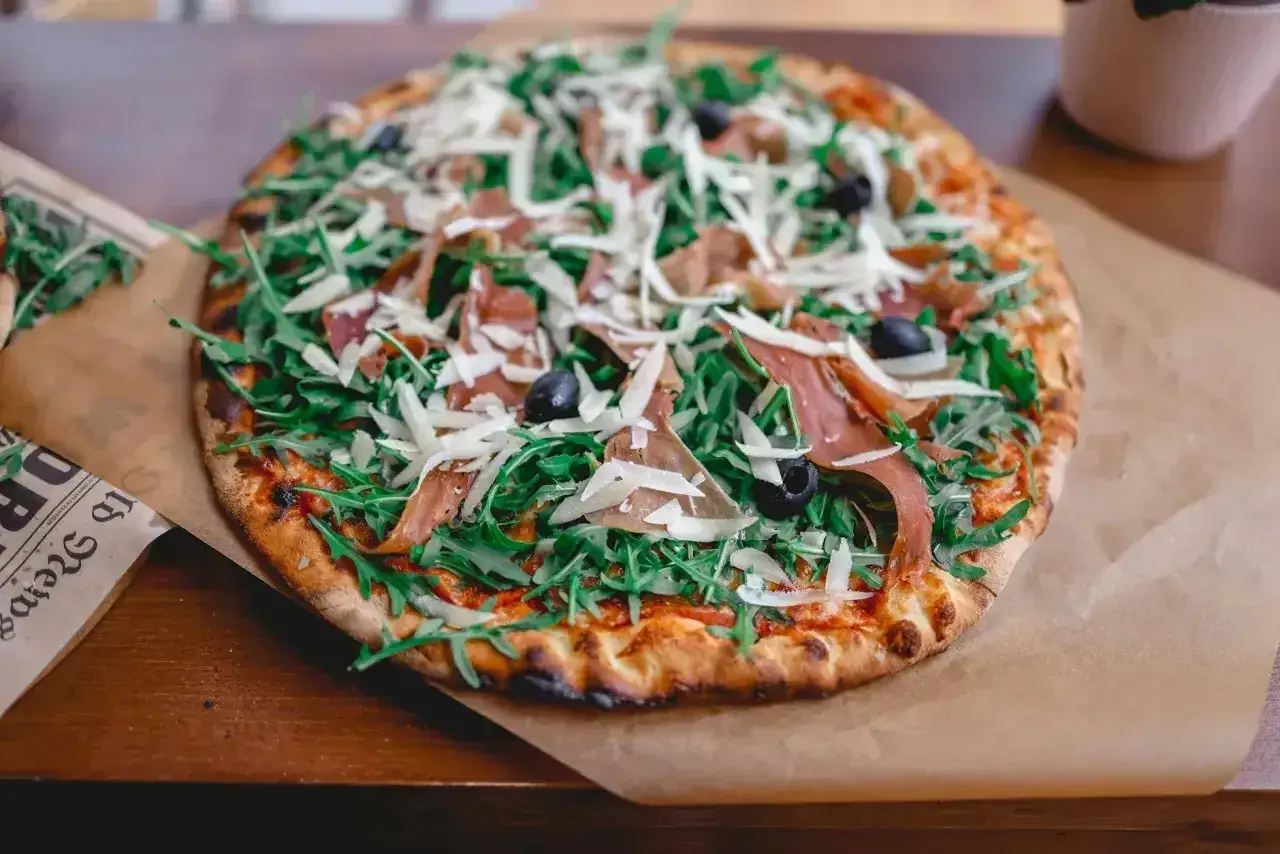 Pyszna pizza prosciutto z rukolą, oliwkami i parmezanem, podana na papierze.
