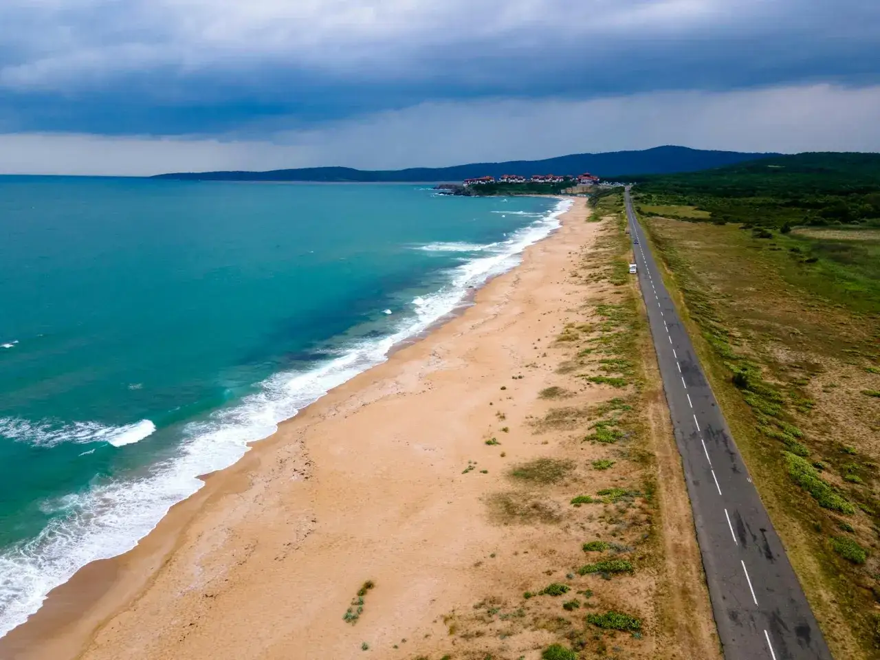 Pusta plaża nad turkusowym morzem, w oddali widać zabudowania i góry. Droga biegnie wzdłuż wybrzeża Bułgarii.
