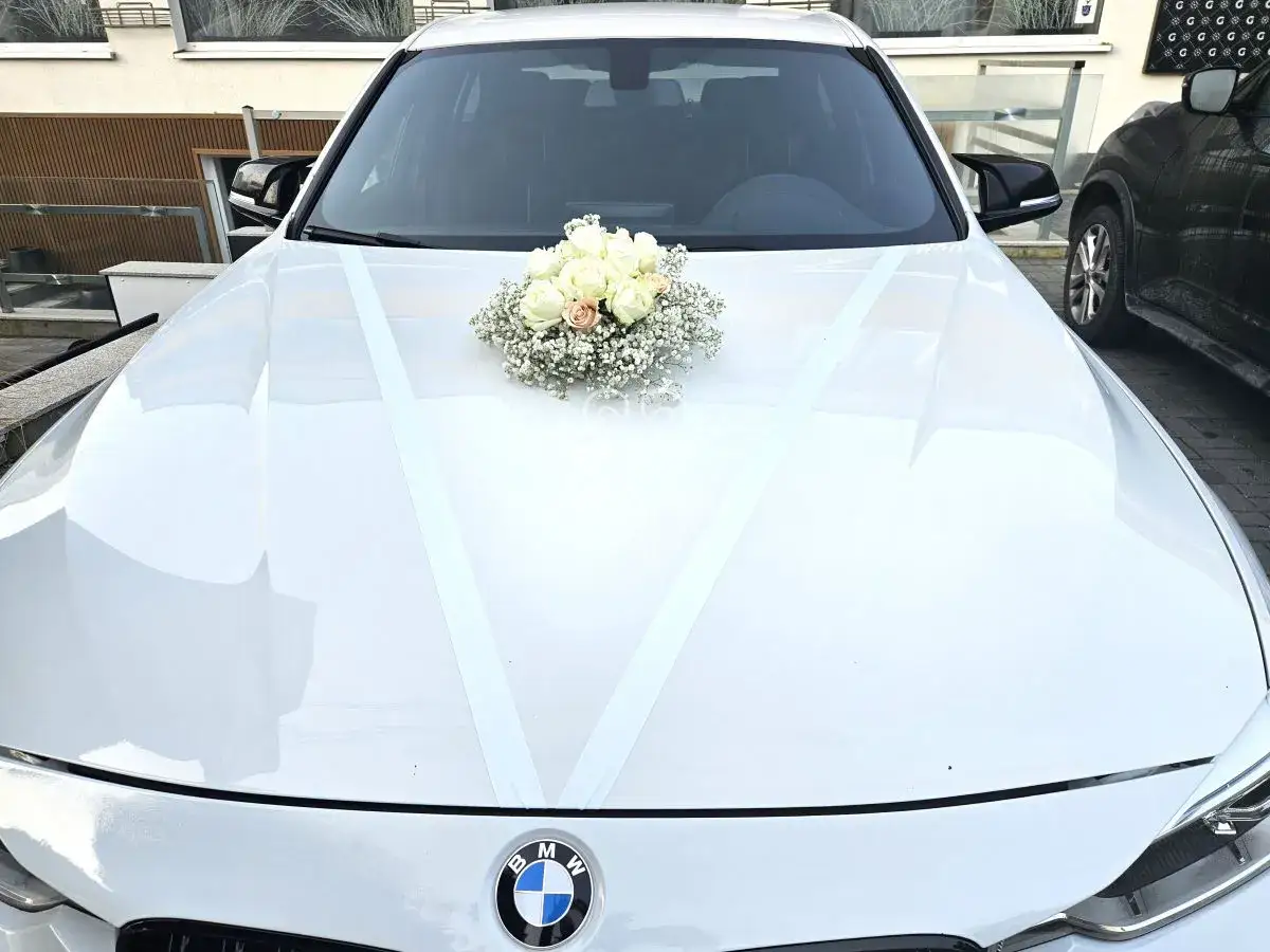 Białe BMW ozdobione wstążkami i bukietem róż, gotowe na ślubną bramę.