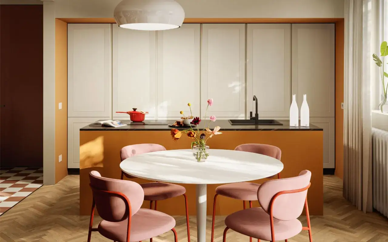 Un tavolo rotondo con sedie rosa accoglienti, un'isola cucina color ocra e un vaso di fiori creano un'atmosfera **clara bona** e invitante.