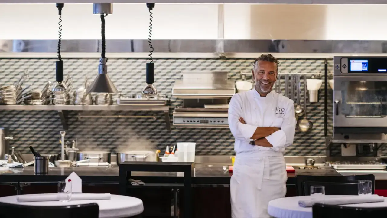 Chef Perbellini, sorridente in cucina, con le braccia conserte. Dietro di lui, attrezzature professionali e tavoli apparecchiati.