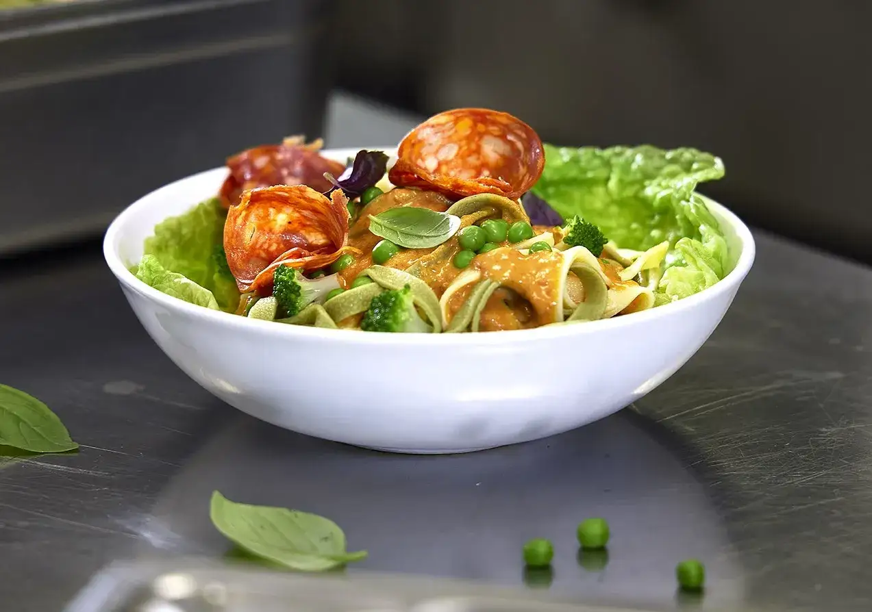 Un plat de pâtes aux légumes colorées, garni de rondelles de chorizo et de feuilles de basilic frais.