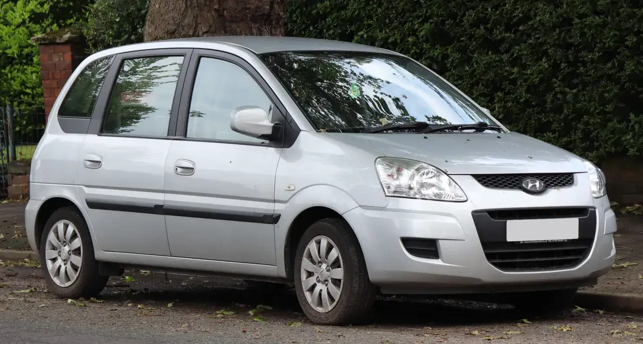 Srebrny Hyundai Matrix zaparkowany przy zarośniętym murku i drzewie.