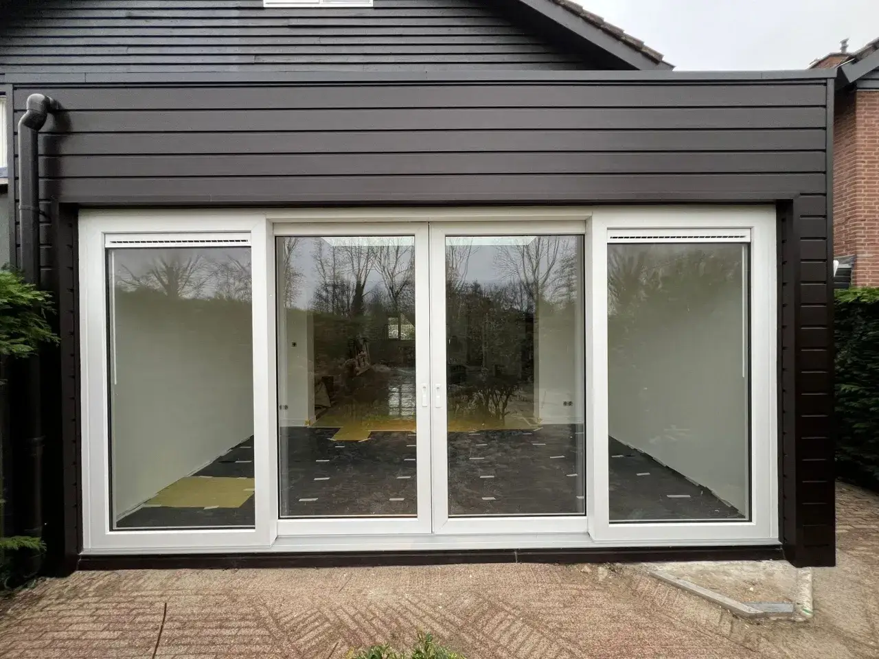 Moderne uitbouw voorkant huis met grote glazen schuifpui. Donkerbruine gevelbekleding en witte kozijnen.