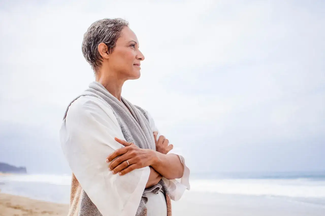 Vrouw met grijze haren op het strand, omarmt zichzelf. Haar blik is naar de horizon gericht, wat haar sensitief betekenis geeft.