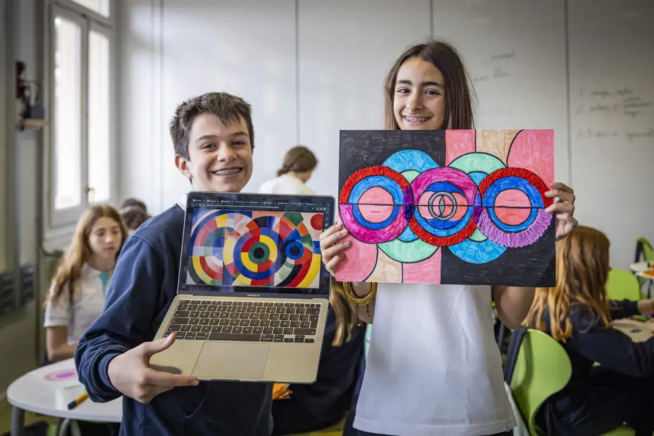 Dos estudiantes muestran sus creaciones artísticas, una en una laptop y otra en papel, como parte de una ia educativa.