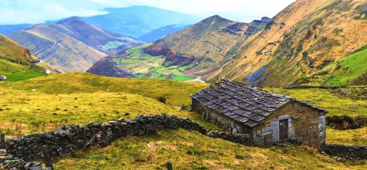 Pequeña cabaña de piedra en los valles pasiegos, rodeada de prados verdes y montañas. Un camino serpentea entre los pueblos.