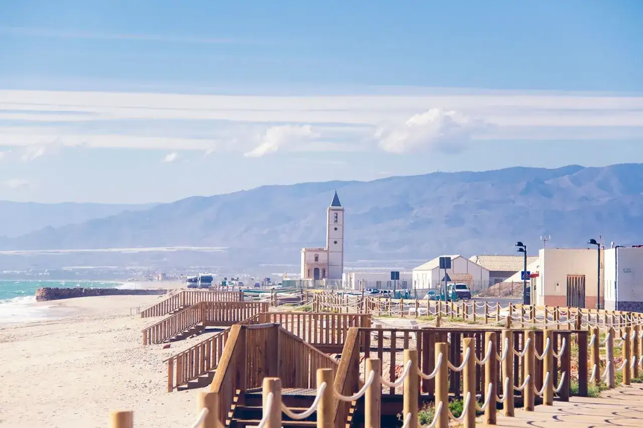 Vista de la playa con escaleras de madera y la torre de la almadraba de Monteleva al fondo, bajo un cielo azul con nubes.