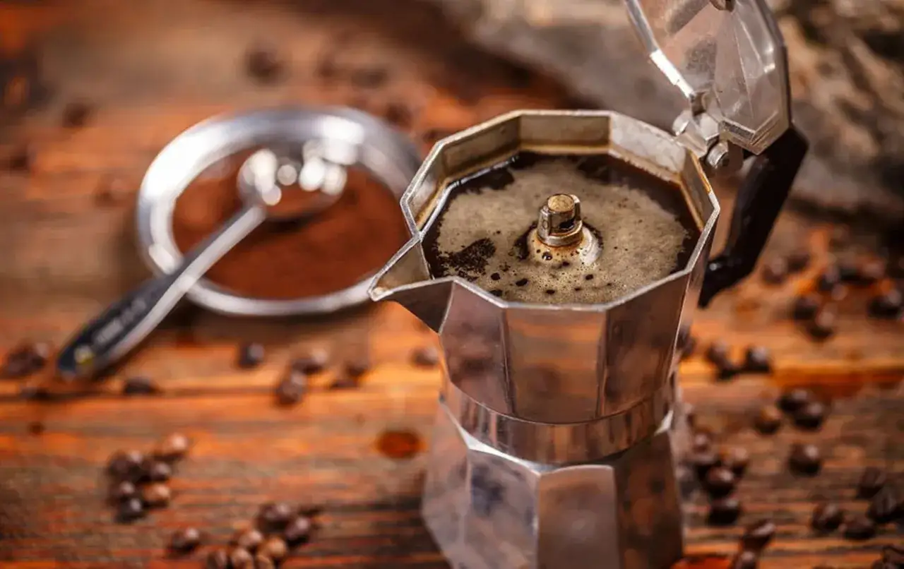 Caffè con la moka fumante, chicchi sparsi su un tavolo di legno. Un cucchiaino riposa in un piattino di caffè macinato.