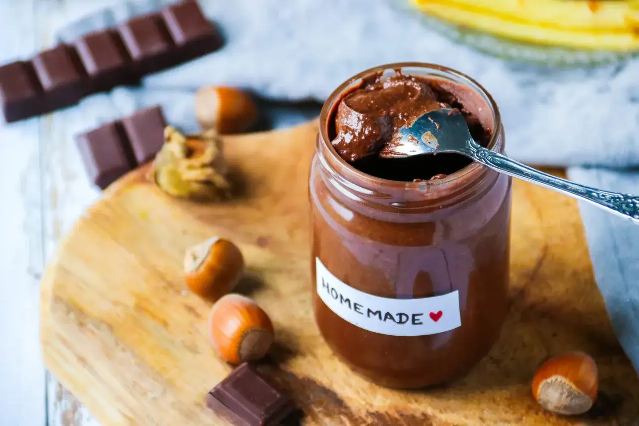 Un pot de p&acirc;te &agrave; tartiner maison 3 ingr&eacute;dients, avec une cuill&egrave;re, des noisettes et des carr&eacute;s de chocolat sur une planche en bois.
