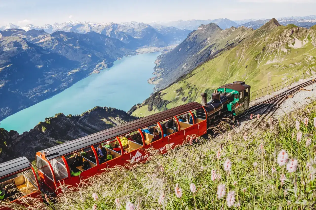 The Brienzer Rothorn train ascends a steep mountain track, offering passengers breathtaking views of a turquoise lake and majestic Alps.