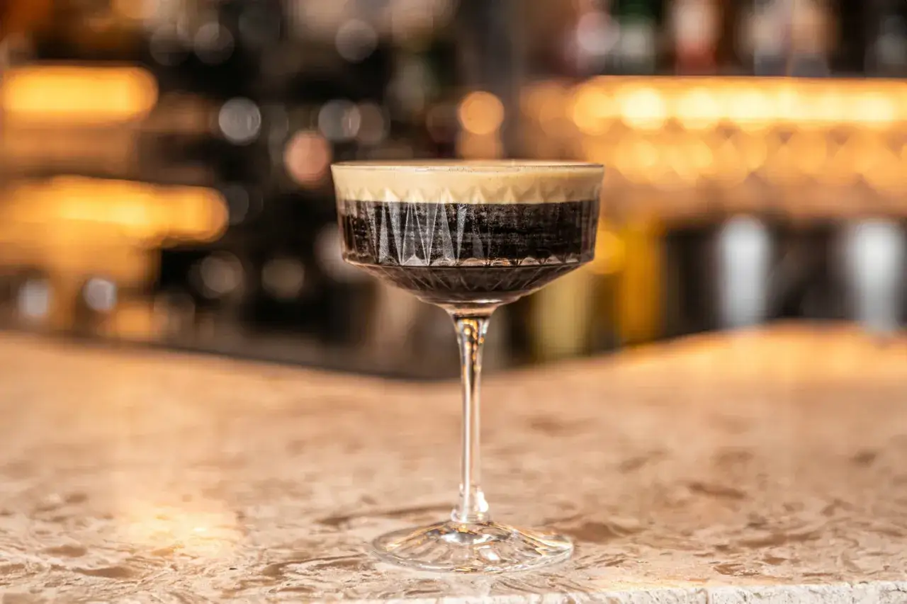 Un elegante bicchiere di Caffe Martini con schiuma cremosa, su un bancone di marmo, con luci soffuse sullo sfondo.