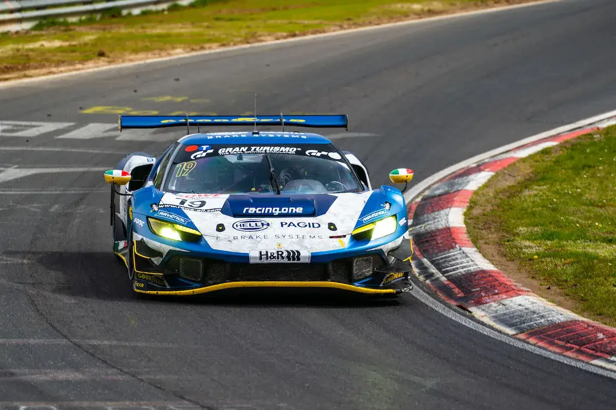 Niebiesko-białe Ferrari 296 GT3 z numerem 19 na torze wyścigowym, z włączonymi światłami.