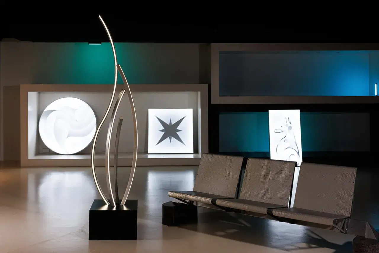 Escultura de luz moderna con curvas elegantes, junto a asientos met&aacute;licos y paneles luminosos con dise&ntilde;os abstractos. La iluminaci&oacute;n crea un ambiente acogedor, con una distribuci&oacute;n de lumenes por m2 que realza cada elemento.