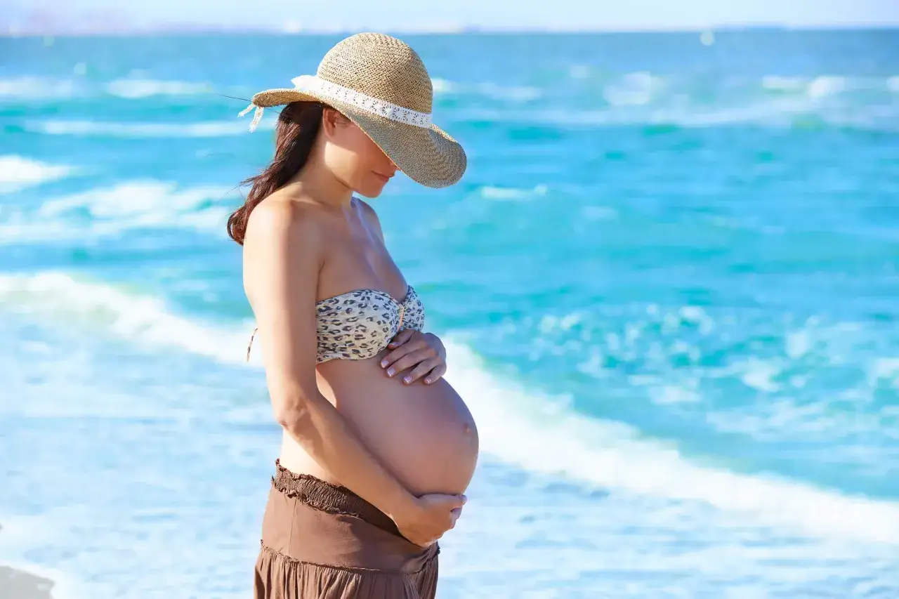 Donna in bikini e cappello di paglia accarezza il pancione, con il mare azzurro sullo sfondo. Un'immagine di un mare in gravidanza sereno.