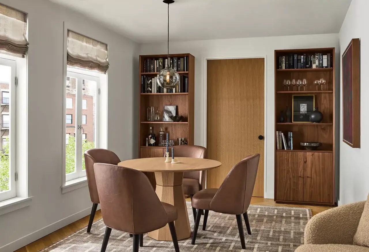 A dining room with a round wooden table, leather chairs, and bookshelves filled with books. The warm wood tones create a cozy atmosphere.