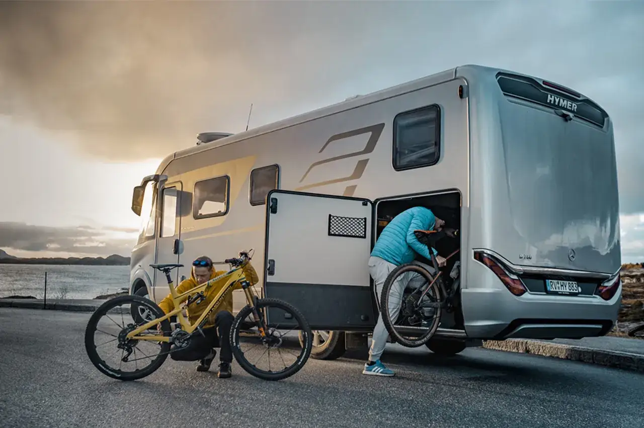 Zwei Personen beladen einen Hymer Wohnwagen mit Fahrrädern. Ein gelbes Mountainbike steht bereit, während das andere e bike im Wohnwagen verstaut wird.