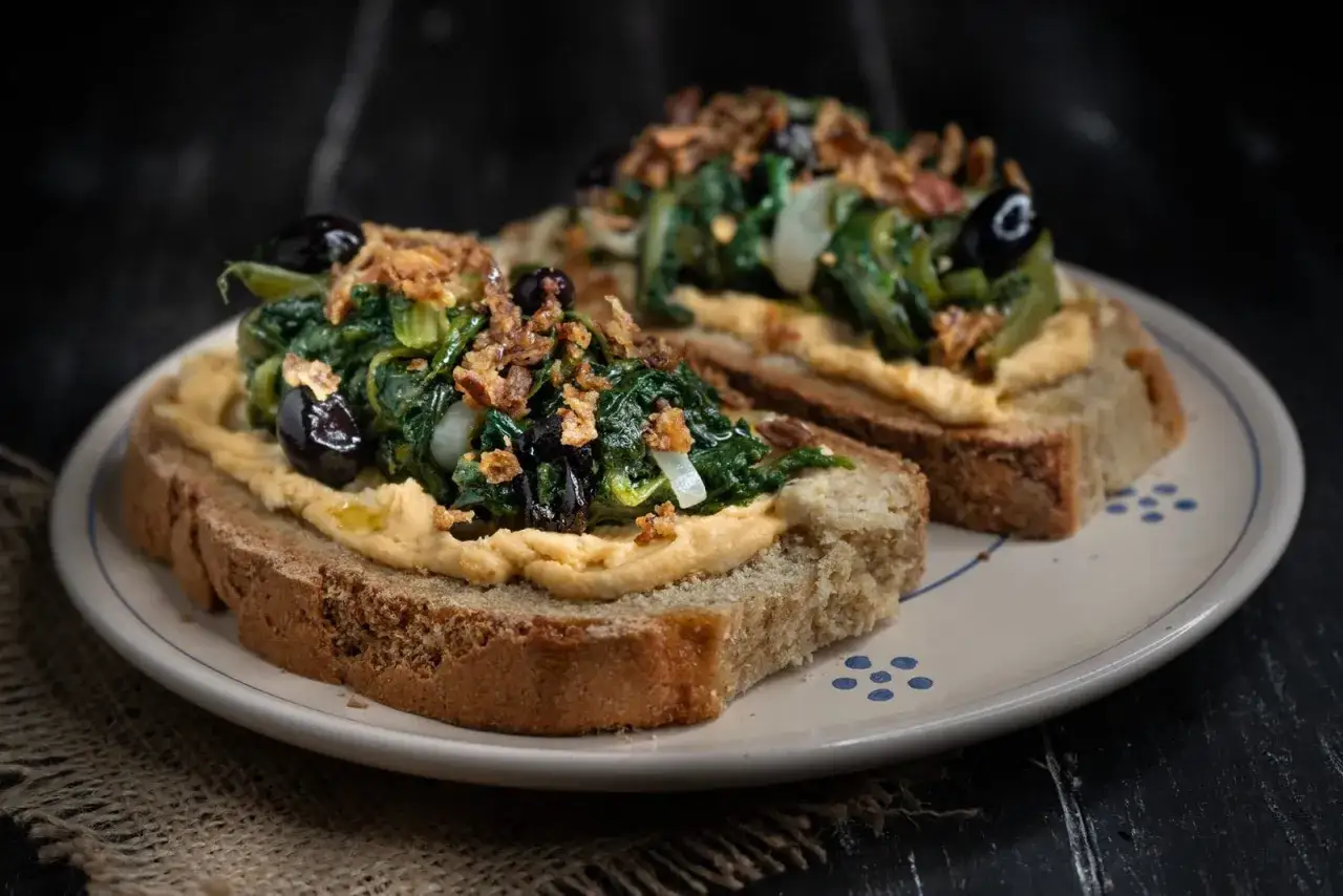Fette di pane tostato con hummus di ceci, verdure saltate, olive nere e cipolla croccante. Un modo gustoso per scoprire come si mangia l'hummus di ceci.