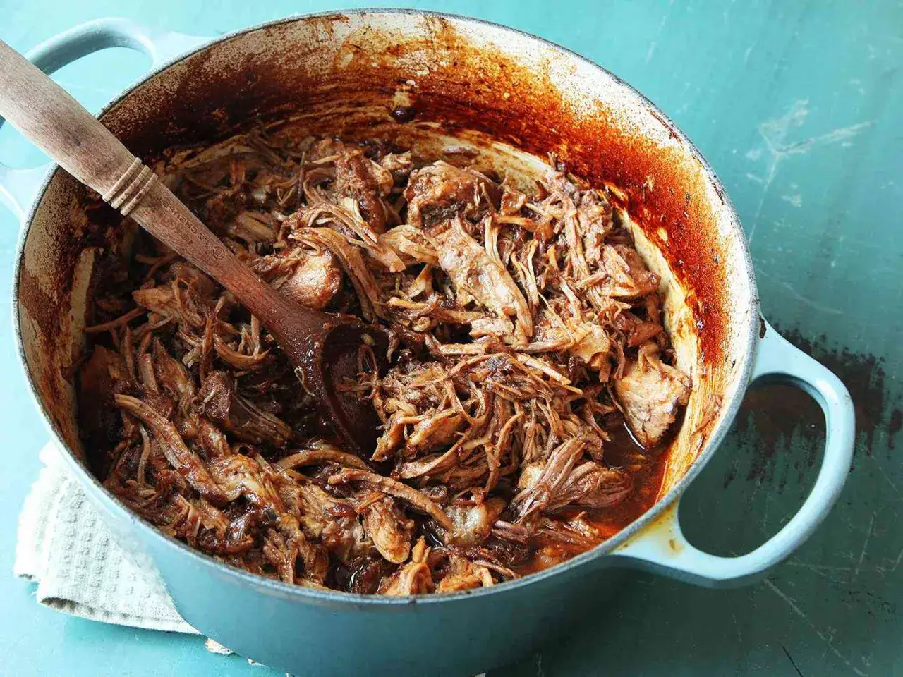 Saftigt pulled pork i ugn, redo att serveras ur en bl&aring; gryta med en tr&auml;sked.