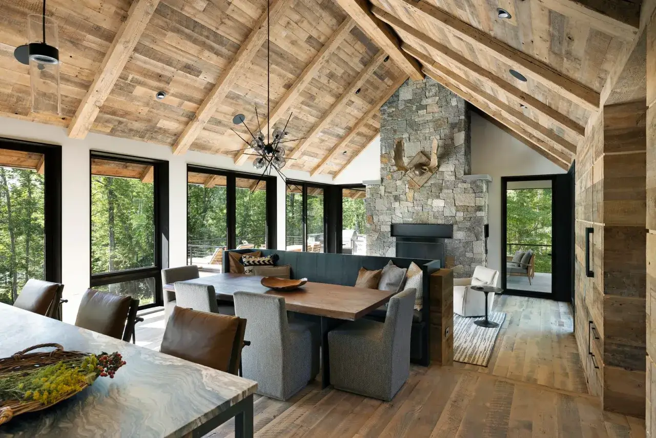 A cozy dining area with a stone fireplace, wooden beams, and large windows showcasing a forest view, embodying traditional rustic interior design.