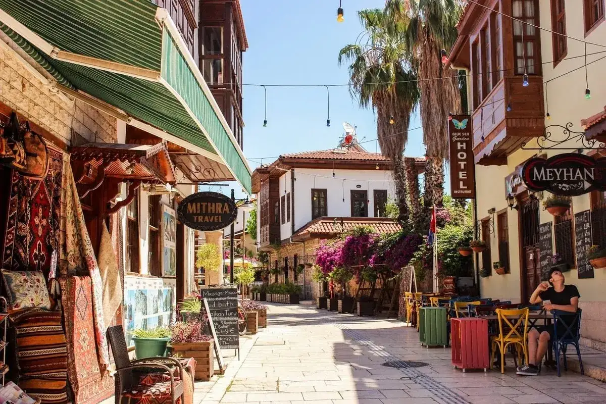 Bunte Gassen in der antalya altstadt mit Teppichläden, Palmen und einem Café, wo ein Mann entspannt sitzt.