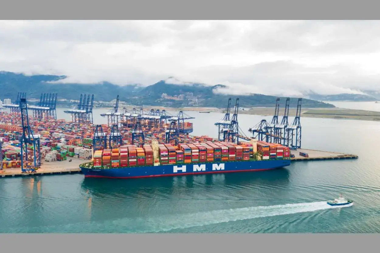 Ein riesiges Containerschiff der HMM-Reederei liegt im Hafen, beladen mit unz&auml;hligen Frachtcontainern. Kr&auml;ne ragen in den Himmel, bereit f&uuml;r die n&auml;chste Ladung.