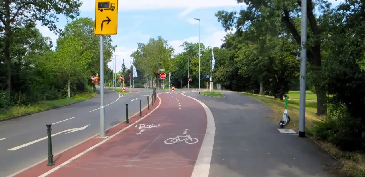 Ein roter, gesch&uuml;tzter Radweg mit Fahrradsymbolen f&uuml;hrt an einer Stra&szlig;e entlang. Ein E-Scooter steht am Wegesrand.