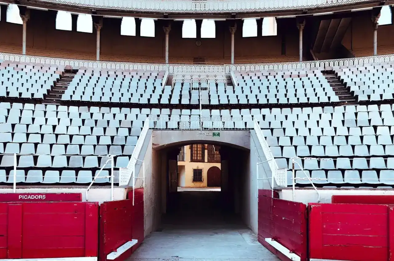 Grada vac&iacute;a de la plaza de toros, con asientos blancos y barreras rojas. Se vislumbra un patio a trav&eacute;s de un t&uacute;nel.