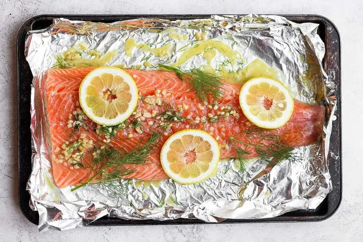 Filete de salmón crudo con rodajas de limón, ajo picado y eneldo sobre papel de aluminio en una bandeja. Listo para cocinar salmón al microondas.