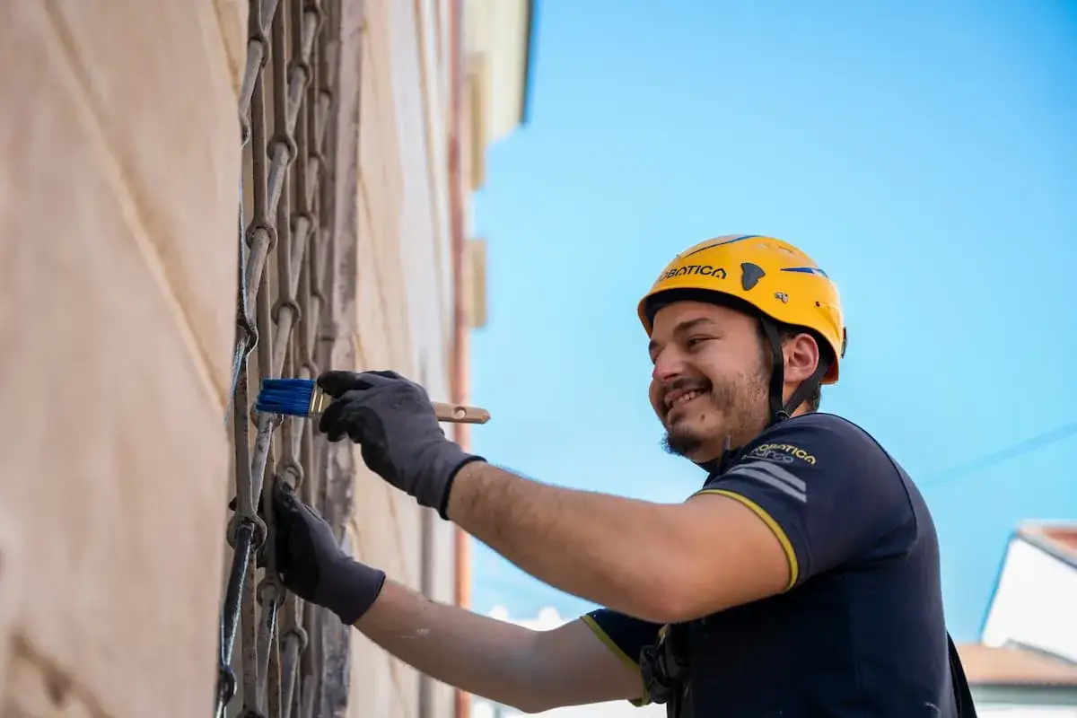 Operaio con casco giallo applica il rasante su una grata metallica con un pennello blu.