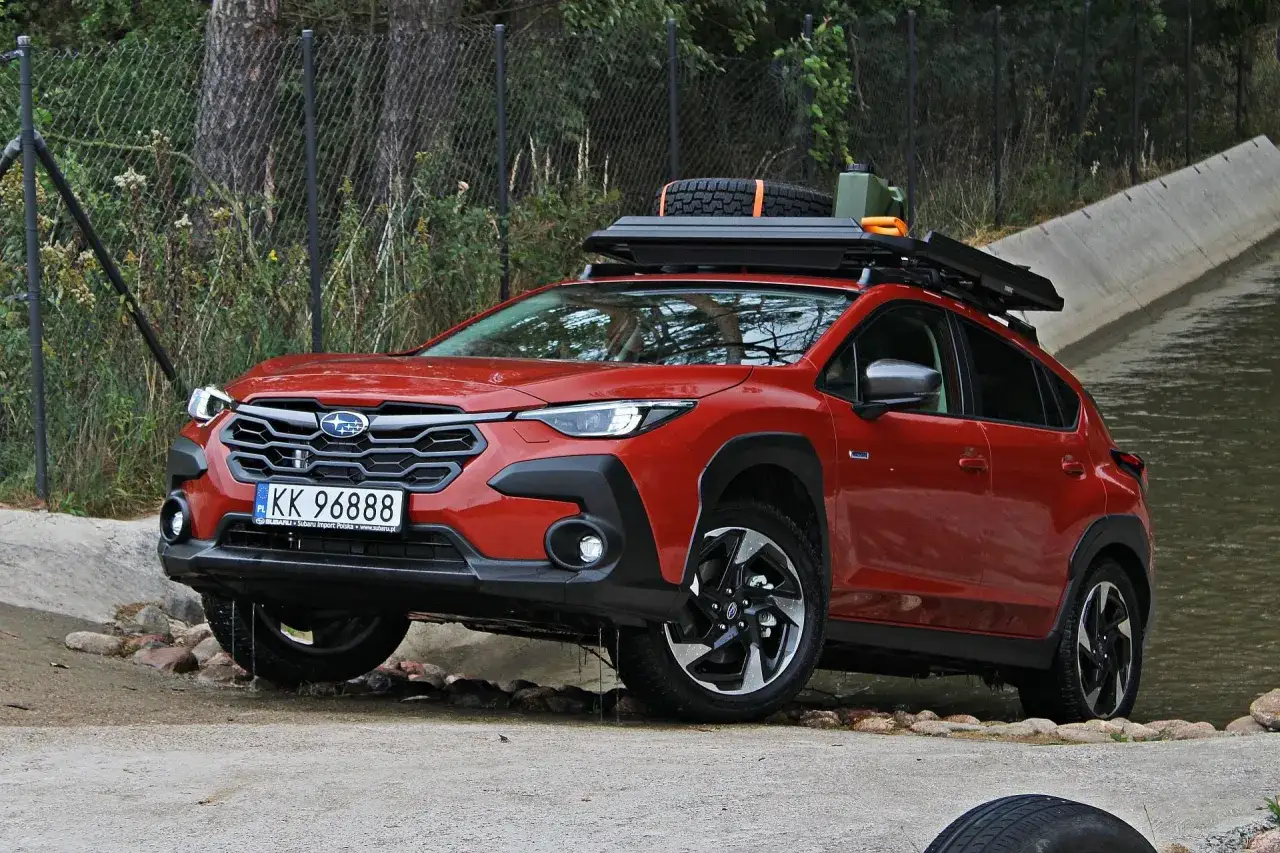 Czerwony Subaru Crosstrek z bagażnikiem dachowym i zapasowym kołem pokonuje przeszkodę wodną.