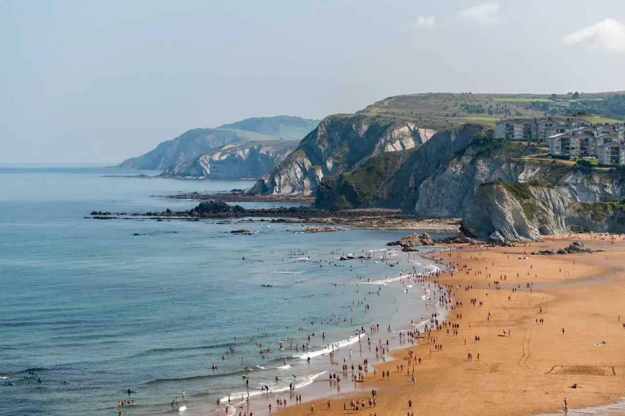 Ein sonniger Tag am Bilbao Meer. Menschen genie&szlig;en den Sandstrand und das erfrischende Wasser, w&auml;hrend sich im Hintergrund steile Felsklippen erheben.