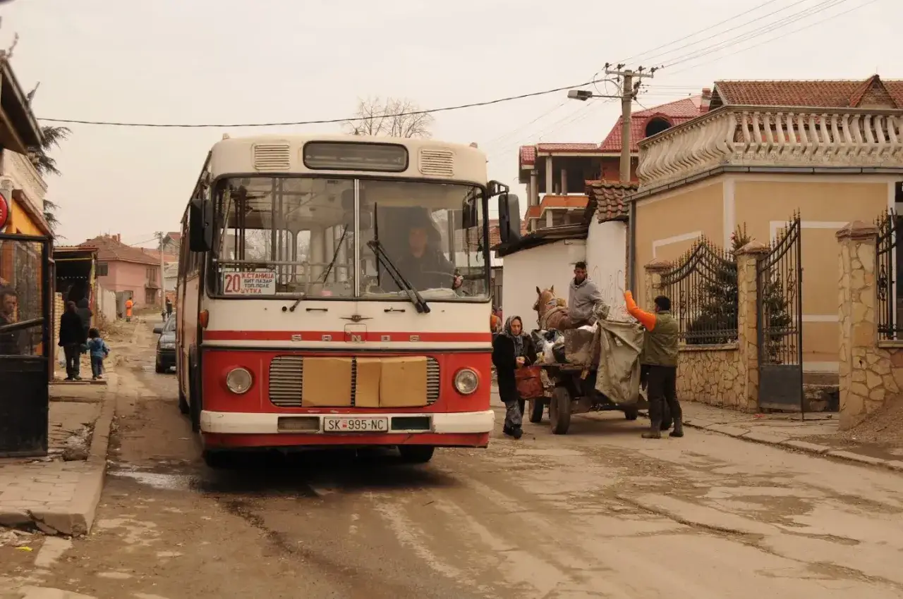 Macedoński autobus z numerem 20 stoi na ulicy. Obok niego wóz zaprzężony w konia, ludzie i domy.