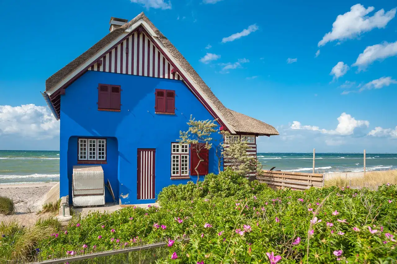 Ein blaues Haus mit Reetdach am Ostseestrand. Perfekt f&uuml;r einen Tagesausflug an die Ostsee, mit Rosenb&uuml;schen im Vordergrund und Wellen im Hintergrund.