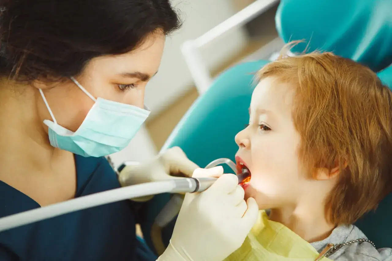 Dentystka bada zęby dziecka, które mogą mieć próchnicę. Mały pacjent siedzi na fotelu dentystycznym, a lekarz używa wiertła.