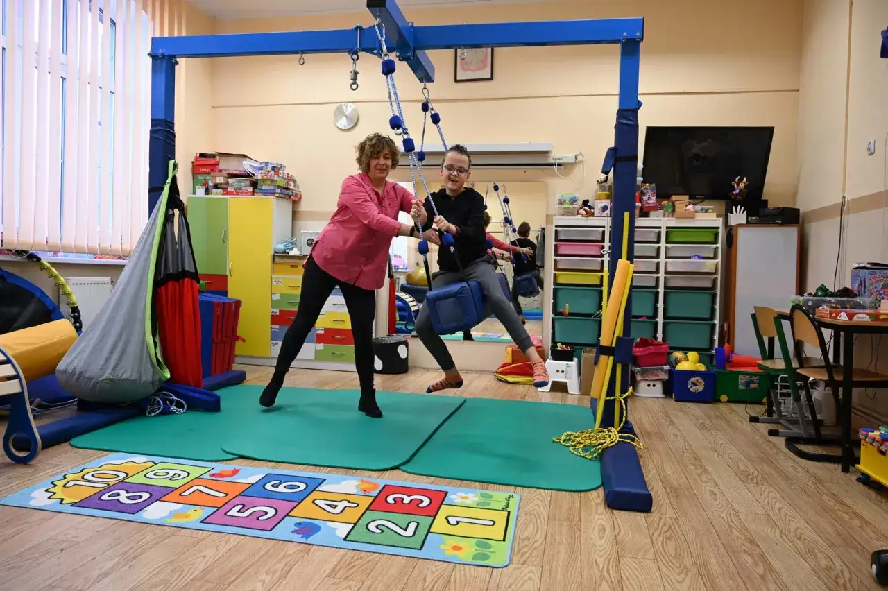 Terapia sensoryczna na czym polega? Dziecko na huśtawce z terapeutką w sali pełnej kolorowych pomocy.