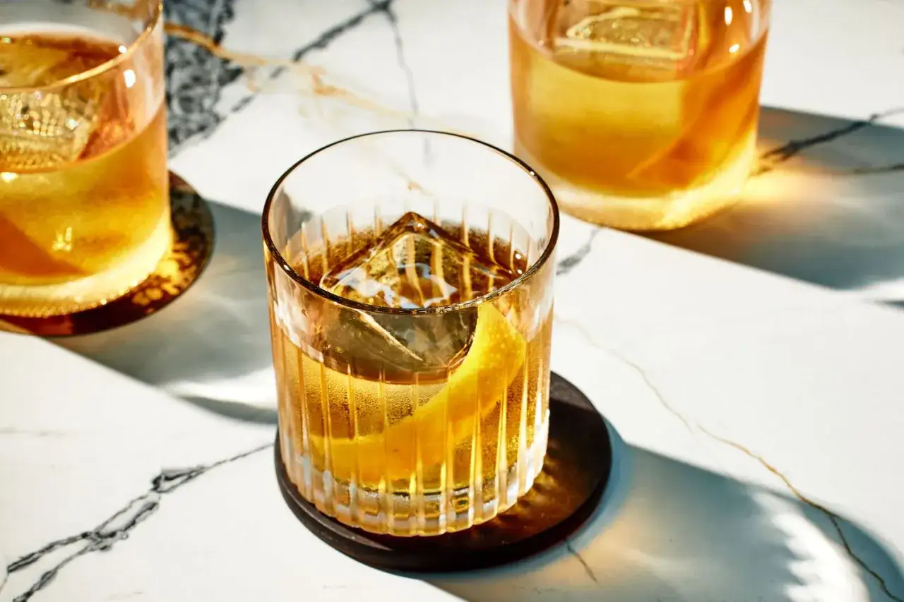 Drei klassische Whiskey-Cocktails mit gro&szlig;en Eisw&uuml;rfeln und Zitronenzesten auf Marmortisch.
