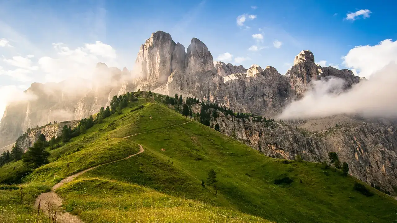 Sentiero tortuoso su un prato verdeggiante ai piedi delle maestose vette del gruppo Sella Dolomiti, con nuvole che accarezzano le rocce.