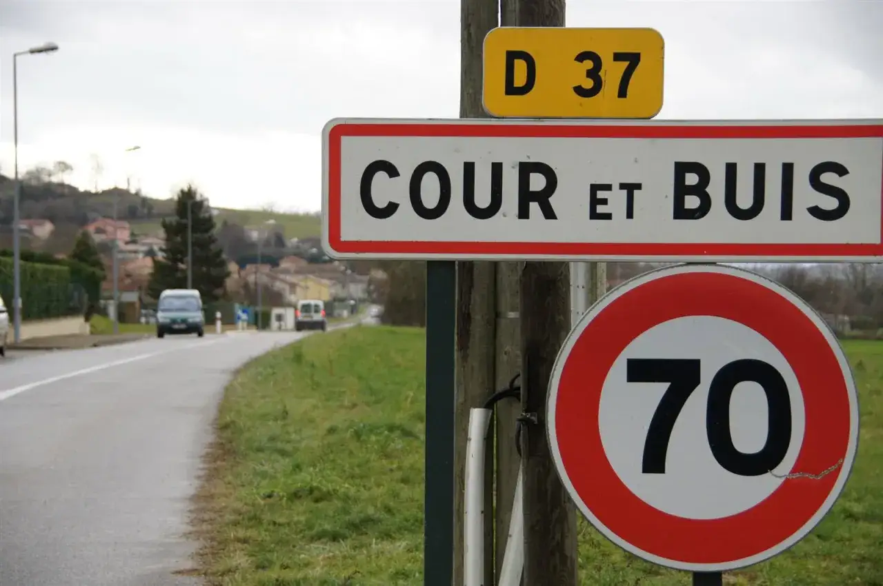 Panneau d'entr&eacute;e de Cour et Buis, avec indication de la vitesse max agglomeration de 70 km/h. Route et maisons en arri&egrave;re-plan.